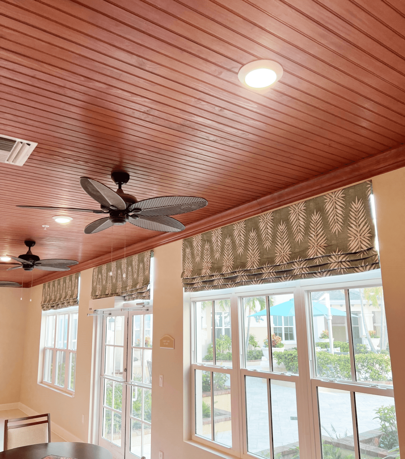 Interior with wooden ceiling, decorative ceiling fans, large windows with patterned curtains, and an outdoor view of a garden with umbrellas and greenery.
