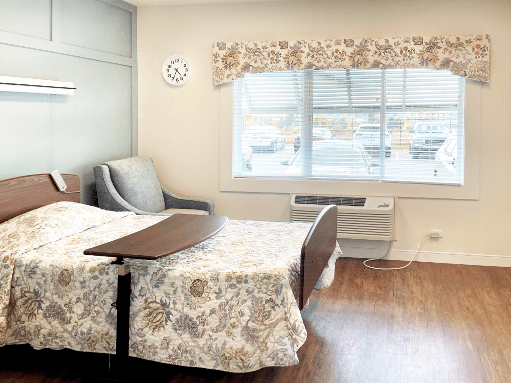 A cozy room with a single bed, floral bedspread, armchair, clock, window, and a wooden floor. Window reveals parked cars outside.