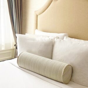 A neatly made bed with a large, rounded bolster pillow, white sheets, and a beige headboard against a softly lit backdrop.