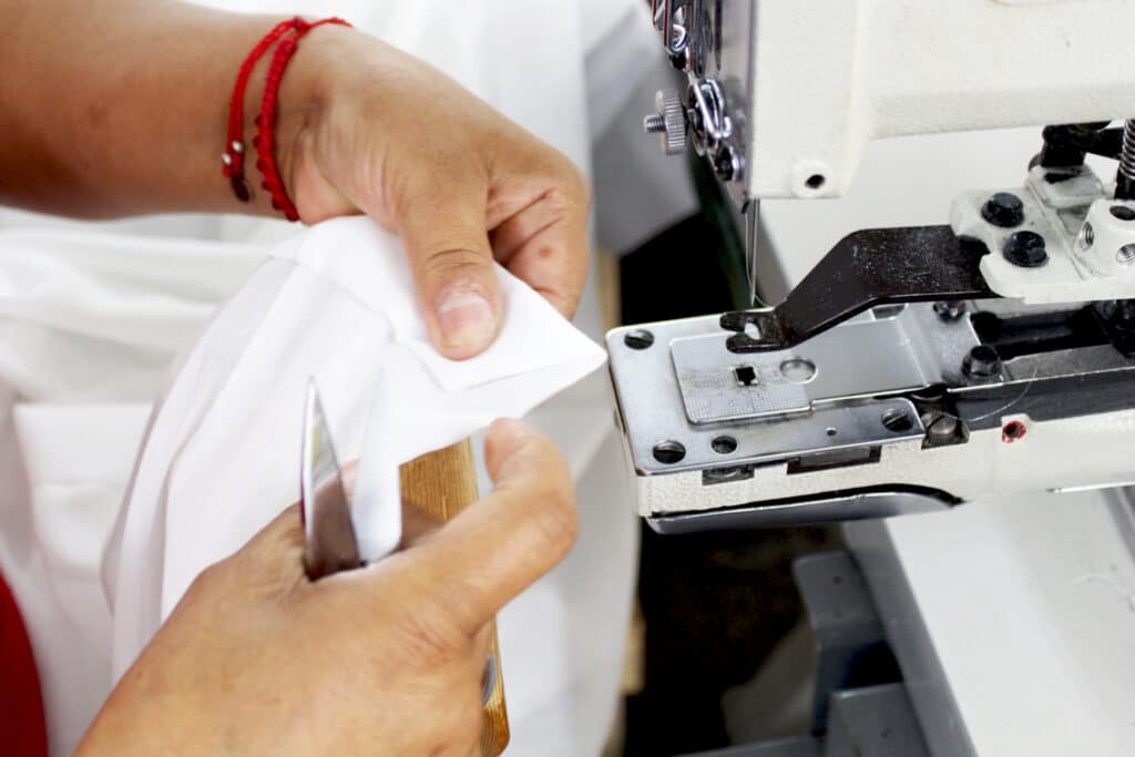 A person sewing white fabric using a sewing machine, with hands visible, skillfully guiding the material, indicating craftsmanship and precision in tailoring.