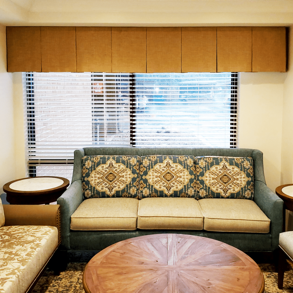 A cozy living room features a patterned sofa, round wooden coffee table, side tables, and window with blinds, creating a warm and inviting atmosphere.