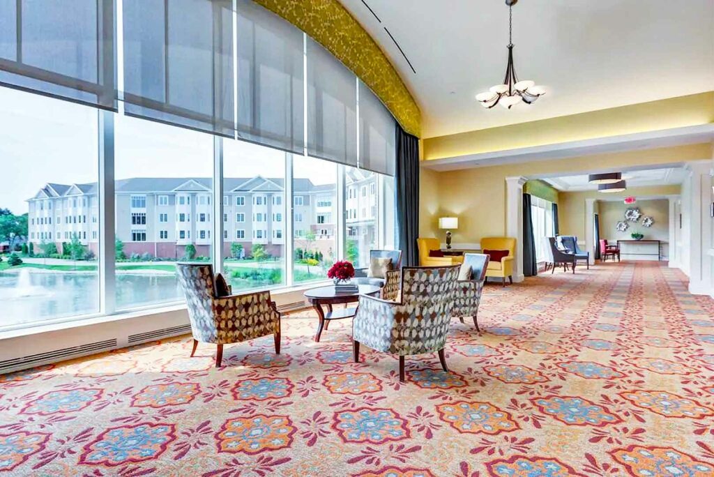 Spacious lounge with patterned carpet, armchairs, and large windows overlooking a pond. Residential buildings visible outside the elegant interior.