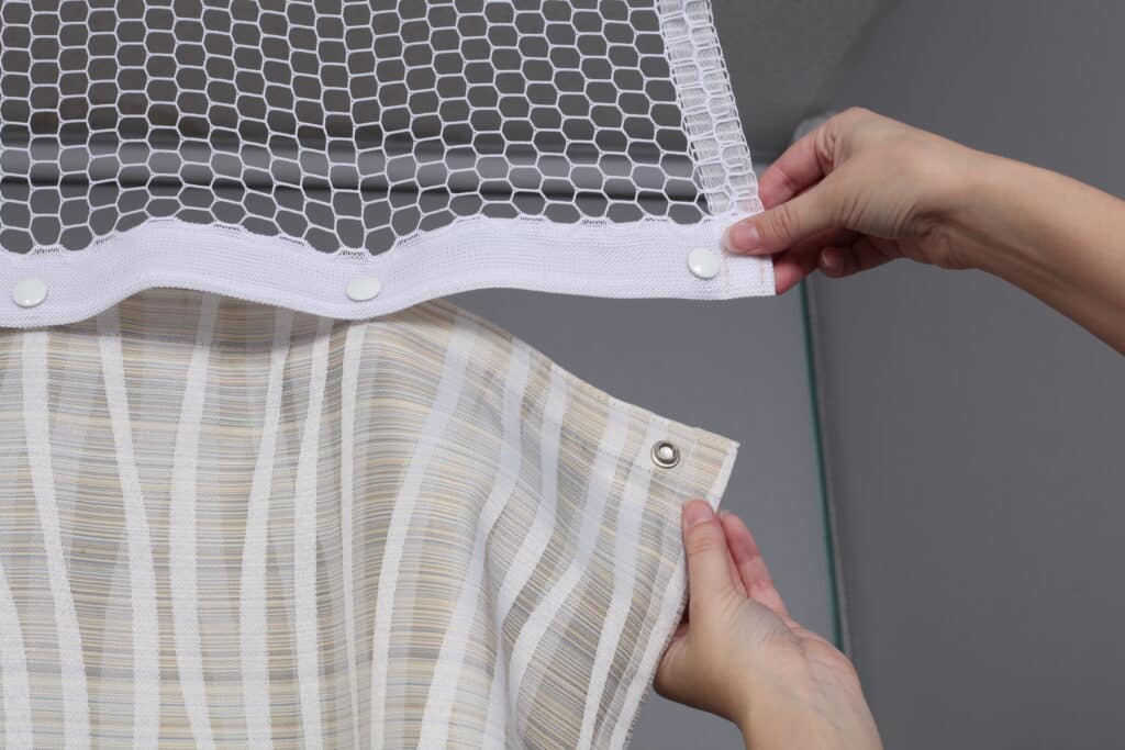 A person is connecting a fabric curtain to a mesh panel with button fasteners, demonstrating assembly or installation of a window treatment.