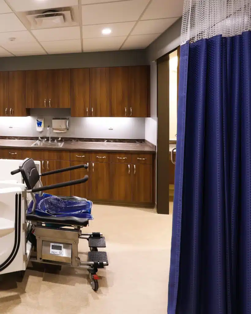 A hospital room with wooden cabinets, a medical chair, and a blue privacy curtain. The space is clean, functional, and well-lit.