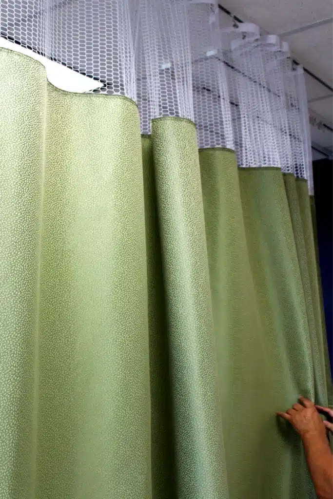 A person adjusts green privacy curtains hanging from a ceiling track, featuring a mesh top for airflow in a healthcare setting.