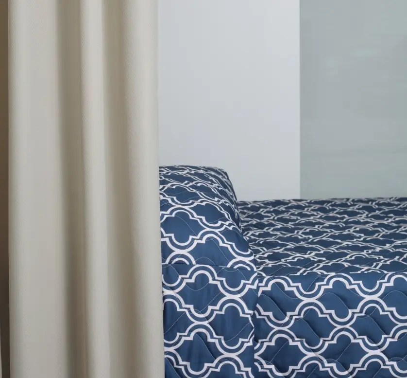 Blue patterned bedding on a bed, partially obscured by a beige curtain, in a minimalistic room with plain walls.