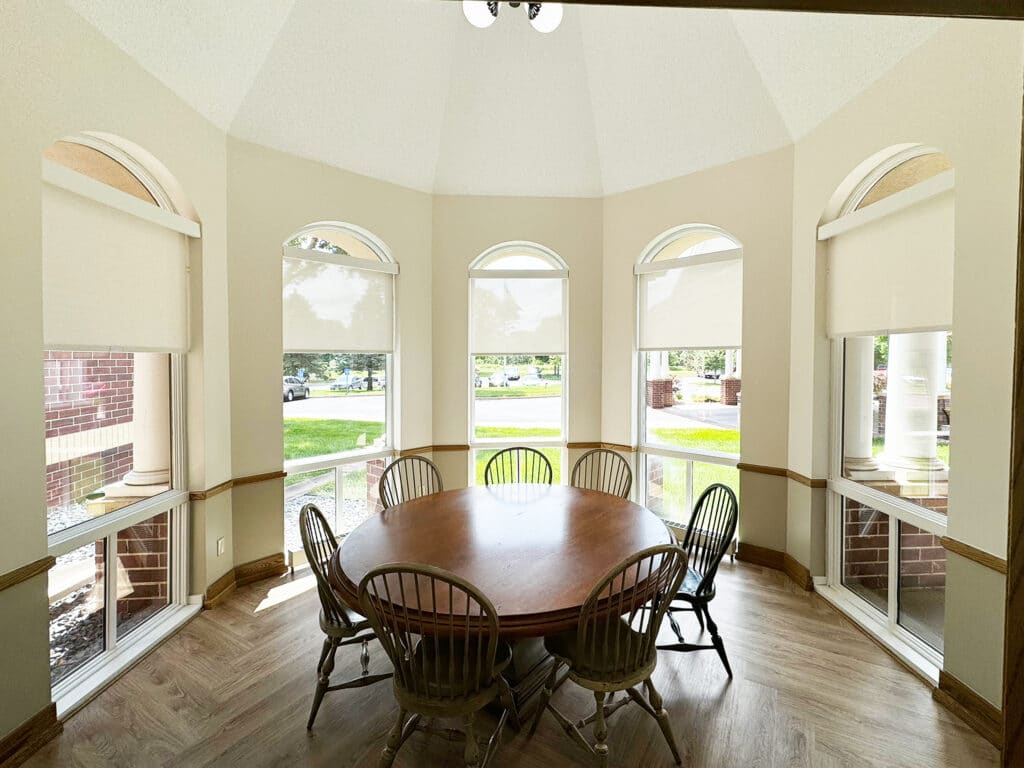 A bright, cozy bay window area with a round wooden table and six chairs, surrounded by arched windows with partial blinds and outdoor views.