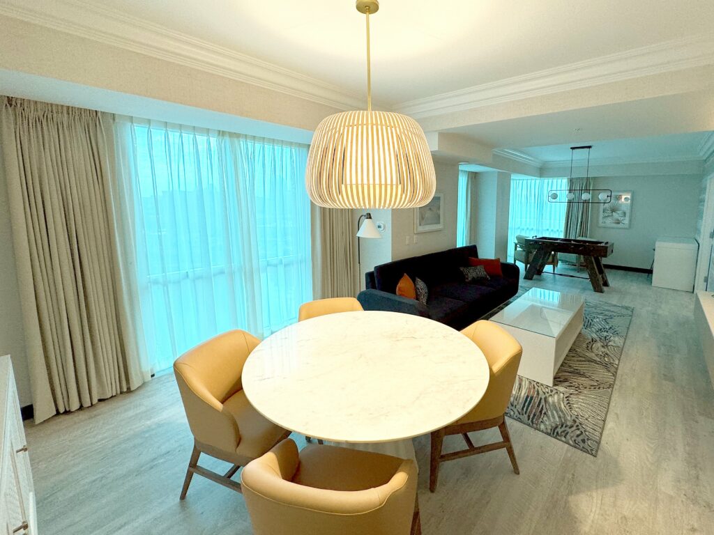 Modern living room with marble table, plush chairs, and a black sofa. Large windows with curtains illuminate the space. Contemporary lighting fixtures.