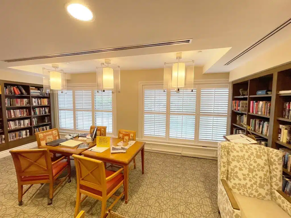 Cozy library with wooden table, four chairs, bookshelves, soft lighting, carpeted floor, and large windows with shutters. Books and papers on table.