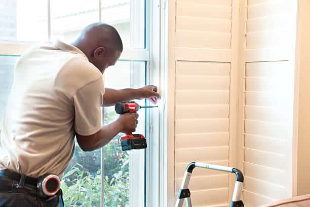 A person installs window shutters using a drill in a bright room. A step ladder is nearby, and there's natural light from outside.