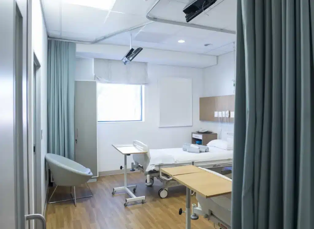 A clean, modern hospital room with a bed, chair, window, and medical equipment. Blue curtains partially frame the scene.