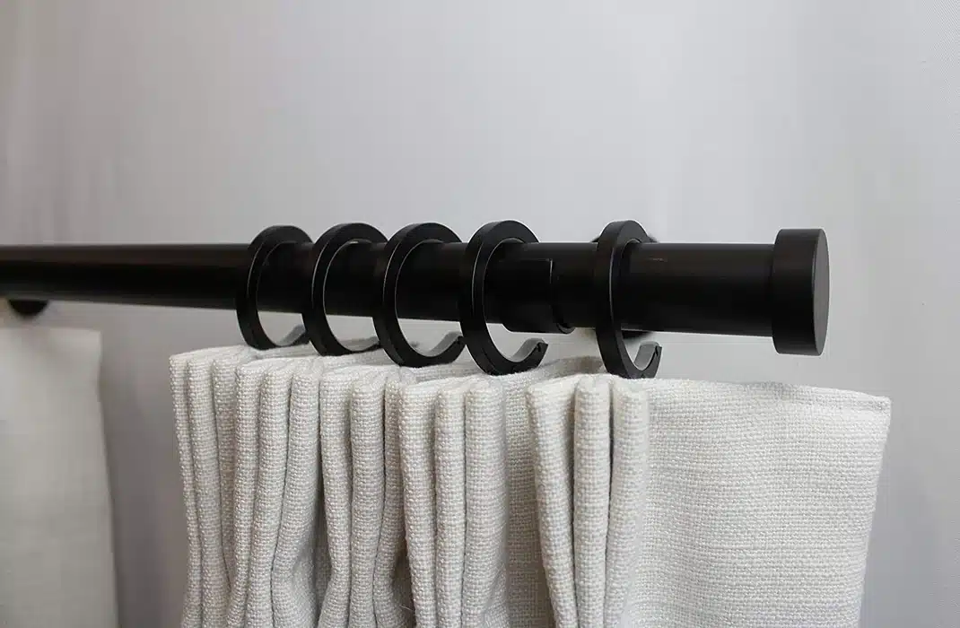 A close-up view of a black curtain rod with white curtains hung using circular rings, set against a plain background.