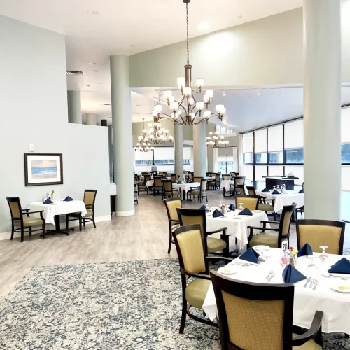 Elegant restaurant interior with neatly set tables, chandeliers, and carpet. Large windows allow natural light. Sophisticated, welcoming atmosphere for dining.