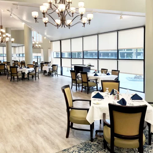 Elegant dining room with chandeliers, neatly set tables, and large windows. The space features light-colored walls and a patterned carpet.