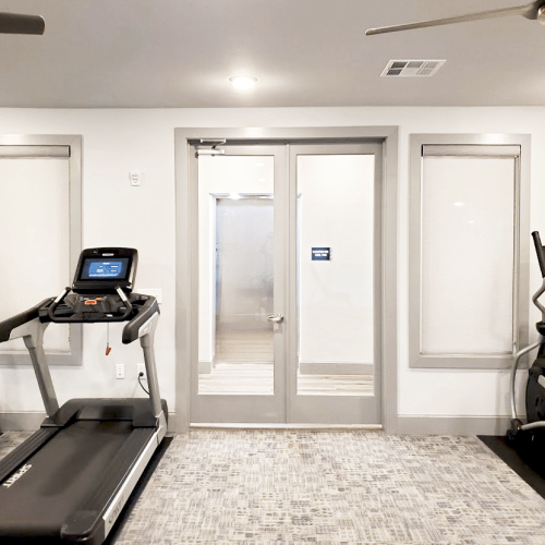 A gym room featuring two treadmills and one elliptical trainer. The room has a neutral color scheme and ceiling fan for ventilation.