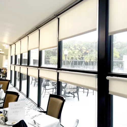 Bright restaurant interior with large windows, blinds, and neatly set tables. Outside, patio furniture and greenery are partially visible through glass.