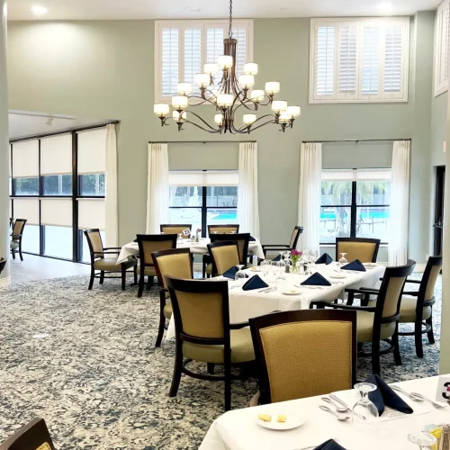 Elegant dining area with neatly set tables, upholstered chairs, and a chandelier. Large windows provide natural light, enhancing the sophisticated ambiance.