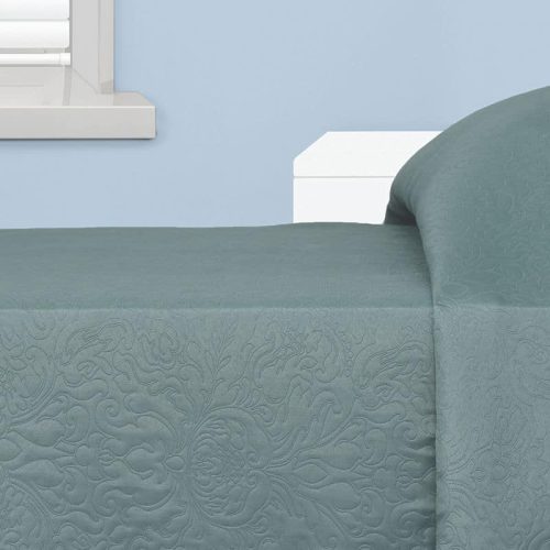 A neatly made bed with a patterned teal quilt is next to a white window with blinds, set against a light blue wall.