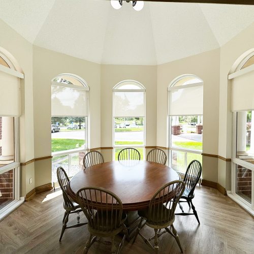 A bright, cozy bay window area with a round wooden table and six chairs, surrounded by arched windows with partial blinds and outdoor views.