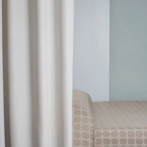 A hospital room with a patterned bedspread, light gray wall, and white curtain. No visible people or specific landmarks present.