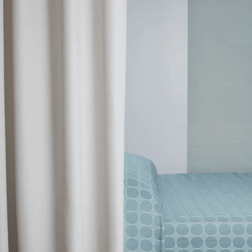 A hospital room with a light blue polka dot bedspread, white curtain, and neutral walls, creating a calm and sterile atmosphere.