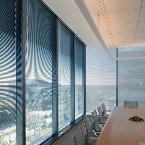 Modern conference room with large windows, overlooking a cityscape. Long wooden table, chairs, and a remote on the table; blinds partially closed.