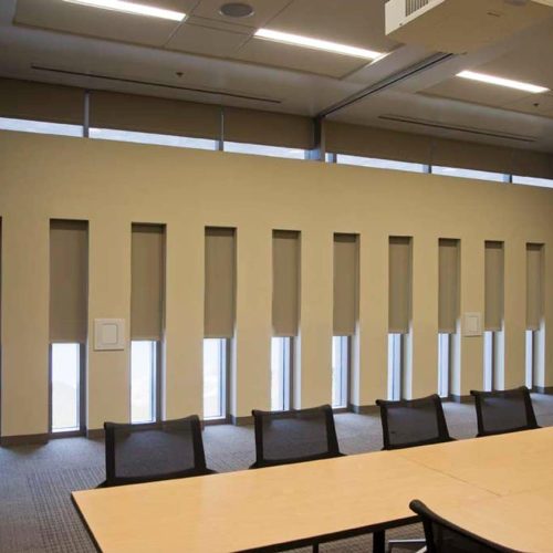 A modern conference room with several tables, black mesh chairs, and vertical window blinds. The walls and ceiling are neutral-colored.