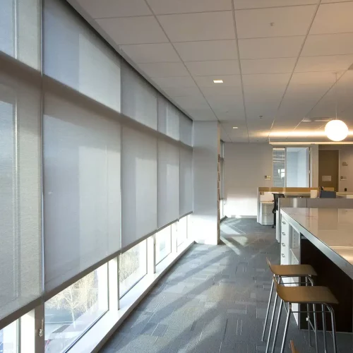 A modern office interior with large windows, blinds, high chairs, and a long counter. Soft lighting creates a calm atmosphere.