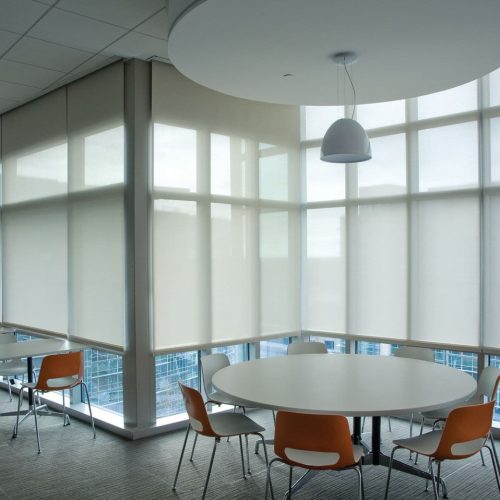 A modern office space with round tables and orange chairs, featuring large windows with blinds and a hanging lamp.