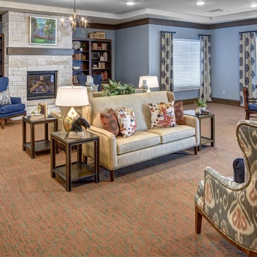 Cozy living room with a stone fireplace, bookshelf, patterned armchairs, and a sofa. Bright windows and decorative pillows enhance the inviting ambiance.