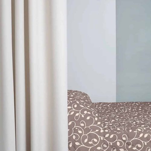 A patterned bed with cream floral design is partially concealed by beige curtains in a minimalist room with light gray walls.