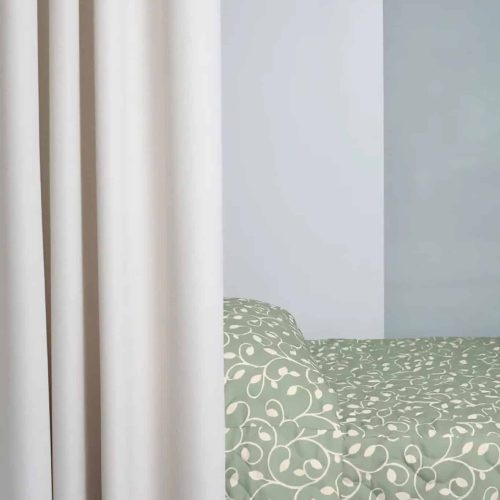 A room with a white curtain partially conceals a green, patterned bedspread on a bed against a light gray wall.