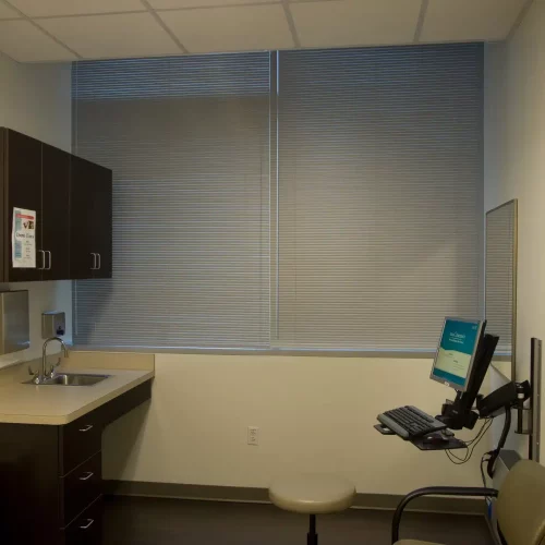 A small, tidy medical office with a sink, computer, and chair. The wall has a landscape painting, and window blinds are closed.