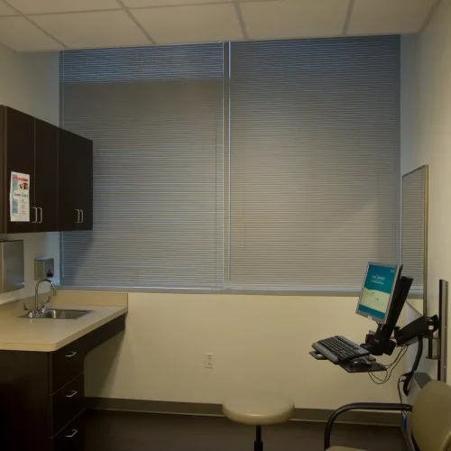 A small, tidy medical office with a sink, computer, and chair. The wall has a landscape painting, and window blinds are closed.