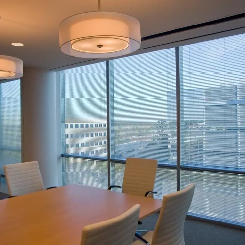 Modern conference room with large windows, overlooking cityscape. White chairs surround a wooden table. Minimalist design with two hanging lamps. No recognizable landmarks visible.