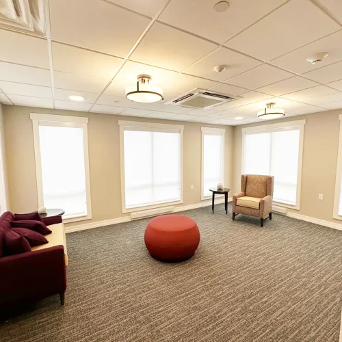 A modern, well-lit room with a maroon sofa, round ottoman, armchair, and several large windows with white blinds.