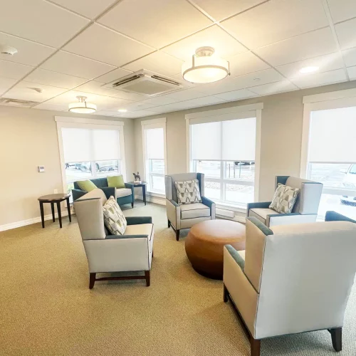 Bright, modern lounge featuring armchairs, round coffee table, and large windows. Neutral colors create a calm atmosphere. No people or landmarks visible.