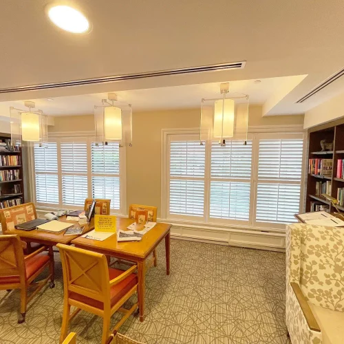 Cozy library with wooden table, four chairs, bookshelves, soft lighting, carpeted floor, and large windows with shutters. Books and papers on table.