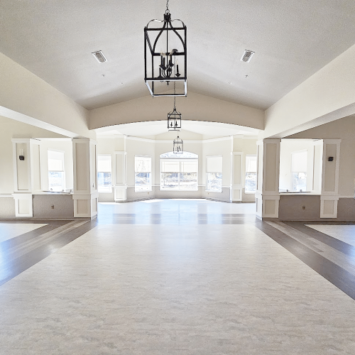 Spacious, elegant room with chandeliers, neutral colors, large windows, and decorative pillars. The well-lit interior features a high ceiling and open layout.