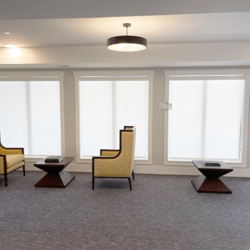 A modern room features three chairs and tables, carpeted floor, and large windows with blinds. Minimalist lighting from ceiling fixture.